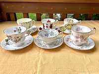 Front view of six complete English teacup and saucer sets arranged on a table outdoors with a wooden fence background. Various floral patterns and gold trim visible.