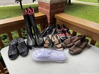 Overview of six pairs of ladies shoes and one pair of disposable slippers on a wooden porch railing and floor outdoors.