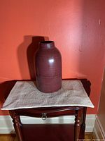 Front view of the burgundy ceramic vase on a small table against an orange wall, showing its shape and color.