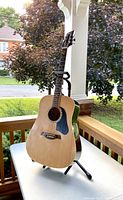 Acoustic guitar with natural wood finish on black stand shown outdoors on table with trees and street background.