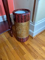 Full view of the signed ceramic floor vase showing maroon body with gold embossed band around the middle featuring equestrian scenes, standing on hardwood floor near wall corner.