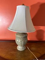 Front full view of lamp with beige ceramic base and white fabric shade on a wooden surface against a red wall.