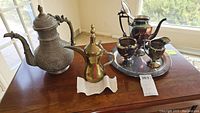 Wide view of all items in the lot on wooden table near window, showing the large brass Arabian Gahwa pot, smaller brass teapot, and silver-toned tea set with tray, pot, creamer, sugar bowl, and cups.