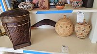 A shelf display showing a tall dark woven lidded basket, two medium woven Southwest style baskets with lids decorated in natural and brown colors with geometric and animal motifs, and five small painted pottery vessels with traditional designs in black and brown on tan.