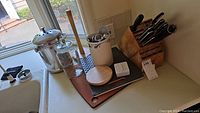 Wide view of wood knife block holding knives, canister, timer, and other kitchen utensils on counter by sink.