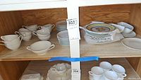 Top shelf wooden cabinet with white cups, saucers, stacks of plates, and small bowls including pieces with decorative yellow and blue rim design.