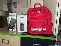 Photo showing the red emergency kit backpack with two boxed items placed beside it on a counter.