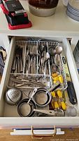 Drawer open showing organized stainless steel flatware in compartment, along with nested stainless steel measuring cups, yellow corn holders, spice infuser, zester, and chopsticks.