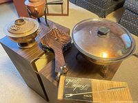 Photo showing three fondue pots: a brass pot with floral lid, a copper pot with spout, and a copper pot with glass lid. Also visible is a wooden paddle and a box of fondue forks.