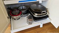 Kitchen utensils including roaster pan, colanders, strainers, baking pans, metal box grater, and wok pan arranged inside a cabinet