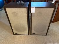 Two vintage Advent LoudSpeakers standing side by side on a carpeted floor, showing front fabric grilles and wood grain veneer sides.