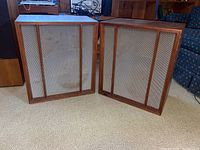 Two vintage wooden speakers with vertical wood bars and metal mesh grill, placed on carpet in living room setting