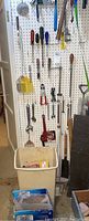 View of a white pegboard wall with various hanging hand tools including screwdrivers, wrenches, pipe wrench, a wire spool on yellow rack, and small shovel tools. White plastic bin and blue cardboard box are placed below with assorted hardware items inside.