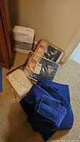 Photo showing white Twin Sheet Set package, two Chattanooga Denim twin duvet covers in plastic packaging, blue towels stacked, and light-colored linen sheet with lace in plastic bag on floor.