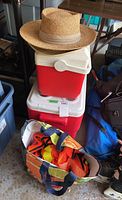 View of two coolers stacked, two natural straw hats on top, and bag of orange safety vests nearby