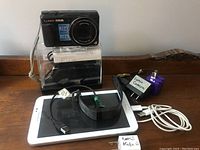 Panasonic Lumix DMC-ZS35 camera showing front with lens and zoom specifications, Samsung tablet, Fitbit fitness tracker, and Altec Bluetooth speaker arranged on table.