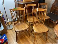 Four armless maple chairs arranged in a room with wooden flooring, showing traditional spindle backs and contoured seats.