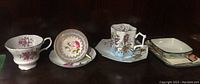 Set of cups and saucers with floral patterns including pink and purple flowers, and gold accents on a dark surface.