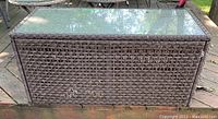 Front angle view of resin wicker chest with clear glass top showing the woven pattern and weathered condition.