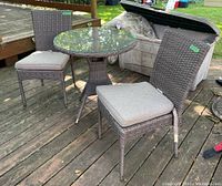 Two resin wicker chairs with cushions and round glass top bistro table on wooden deck outdoors. Weathered condition visible on wicker.