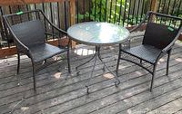 Patio set on wooden deck showing two resin wicker arm chairs and round glass top bistro table.