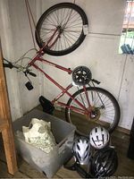 Red Raleigh Ozark 18 speed bicycle next to wall in shed with three helmets on the floor beside it. Clear view of frame, wheels, and helmets.