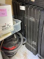 Stacked, taped black plastic shelf unit pieces with vacuum cleaner and storage bin nearby in garage setting.