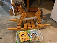 Wooden rocking horse with two children's books placed in a plastic container under it, photographed on concrete basement floor near stairs.