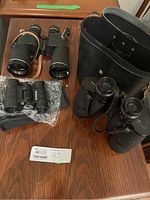 All three binoculars placed on wooden table along with black carrying/storage case and soft pouch