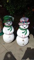 Pair of hand-painted concrete snowmen with green and red hats, standing on outdoor paving.
