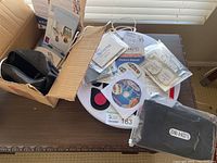 White circular foot massager unit with instruction manuals, adhesive pads, CD, and black leg wrap on table