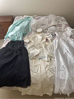 Photo showing full spread of six dresses and veil on bed including light blue dress, black dress, and three white/cream wedding dresses with veil and satin cap.