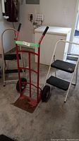 Photo showing red steel hand truck with inflatable tires and two white steel folding step stools with black steps in background.