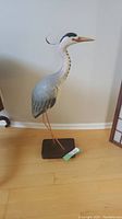 Full side profile of the large standing grey heron sculpture on wooden floor against wall.