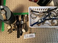 Overview of men's and ladies' watches on floor and in basket; includes metal band watches, Garmin watch, white basket of additional watches