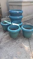 Five blue glazed clay plant pots of varying sizes placed on concrete. Each pot is round with a flared lip.