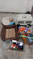 Photo showing two dog beds, one cream round and one brown square, a plastic pet crate, and a container with assorted toys and leashes