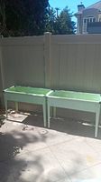 Two green metal raised garden beds side by side shown outdoors on concrete with a privacy fence in the background.