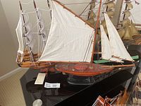 Schooner-style sailing ship model with white sails and reddish-brown wooden hull shown from the side with rigging and deck details visible.