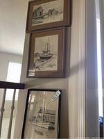 Three framed ink and watercolor drawings hanging vertically and leaning on a wall. Depict boats and waterfront buildings.