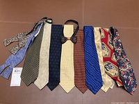 Photo showing 10 silk neckties laid out in a row on a brown surface, showing various patterns and colors, plus one dark leather bow tie with a black strap.
