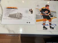 Top down photo of the signed Boston Garden postcard and signed Bobby Orr hockey card laid flat on a white marble surface.