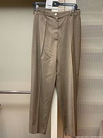 Full front view of beige wool trousers hanging on clip hanger against perforated metal background