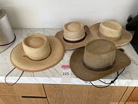 Four cowboy style hats on a marble surface: two tan felt hats with chin straps, one light beige felt hat, and one natural woven straw hat with black band and chin strap.