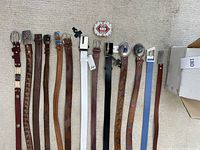 Top view of 11 belts arranged side by side on carpet, showing various colors, buckles, and decorative details