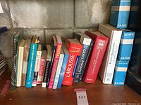 Photo showing multiple reference and photography books arranged on a wooden shelf against a concrete wall. Titles visible include Physicians Desk Reference, Guinness World Records 2013, Larousse dictionary, Gray's Anatomy, and photography books.