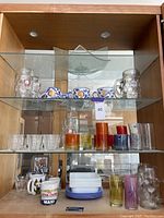 Overview of multiple shelves showing the Italian mugs, beer steins, assorted clear and colored drinking glasses, coffee mugs, and stack of plates and saucers.