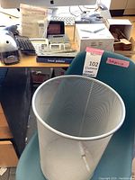 Photo showing mid-century style teal chair and silver metal mesh wastebasket in front of a desk with electronic items