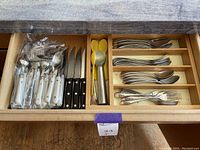 Drawer with assorted utensils including white handled forks, knives and spoons, black handled steak knives, yellow serving spoons, and metal forks and spoons.