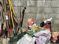 Gardening tools including shovels and green plastic rakes with metal handles leaning against a wall next to multiple bags of soil and fertilizer.